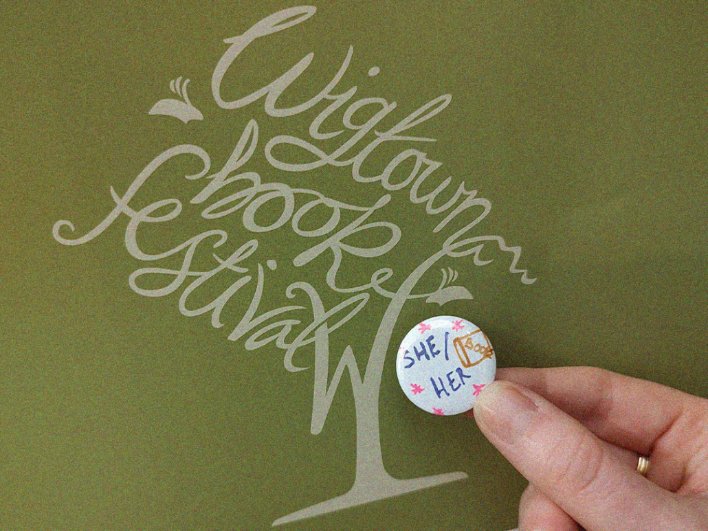 Wigtown Book Festival Logo with a she/her pronoun badge in front of it.